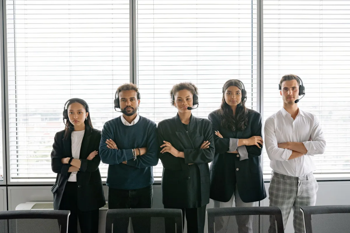 A diverse group of call center agents standing with arms crossed, wearing headsets in an office.