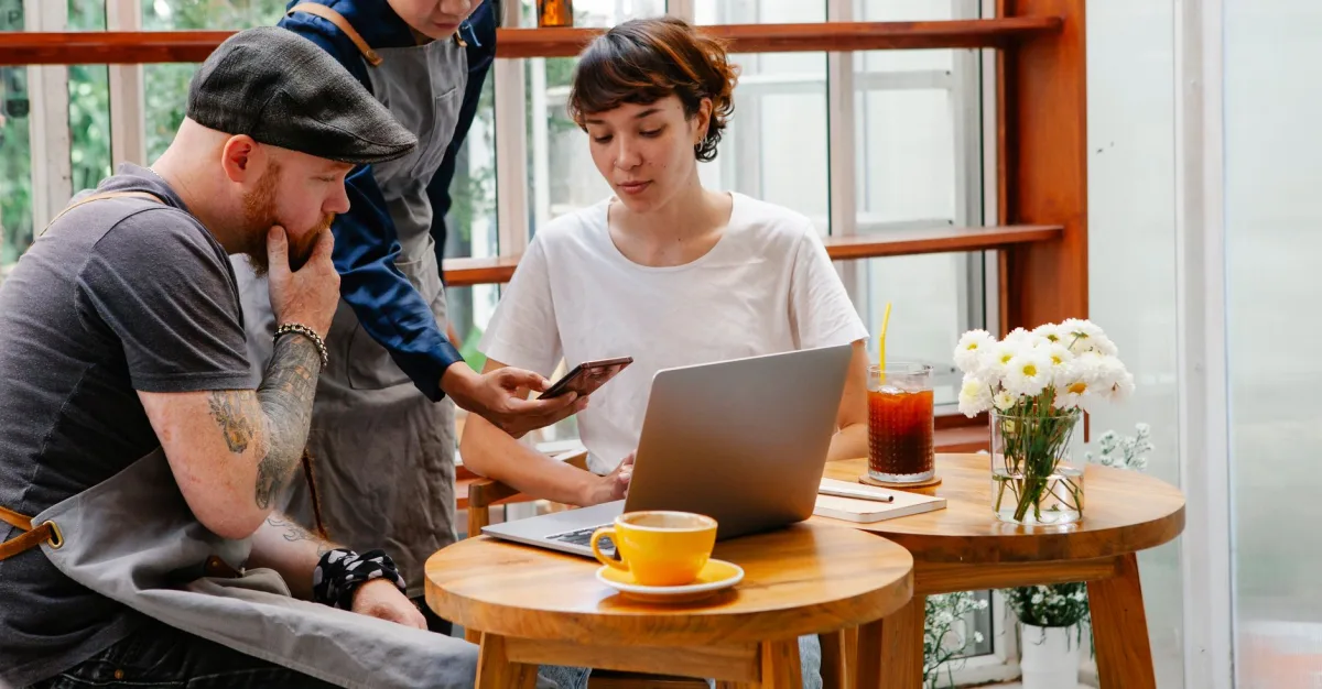 Concentrated coworkers in aprons sitting at table with laptop while looking at smartphone of anonymous colleague in cafeteria during work