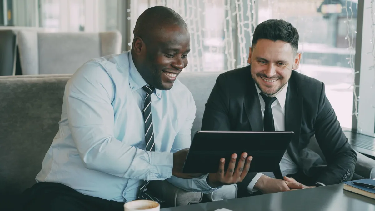 Two businessmen happily collaborating on a tablet in a modern cafe setting.