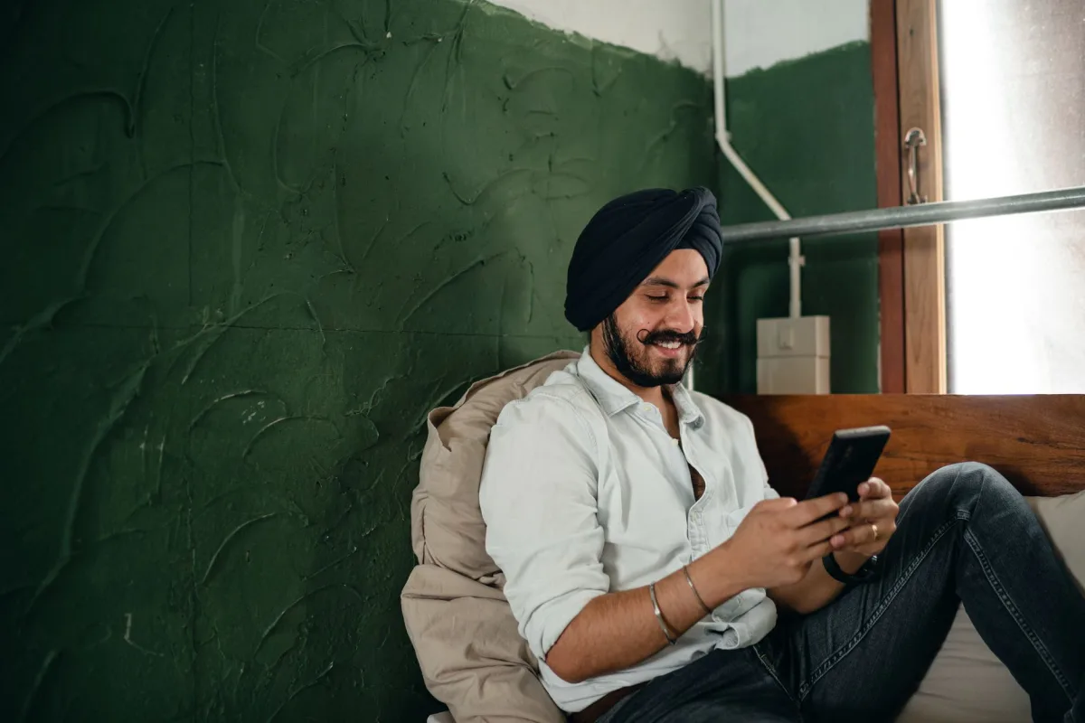 Young bearded Indian male in casual clothes and turban sitting on bed and leaning on pillow against roughly plastered green wall while messaging on mobile phone