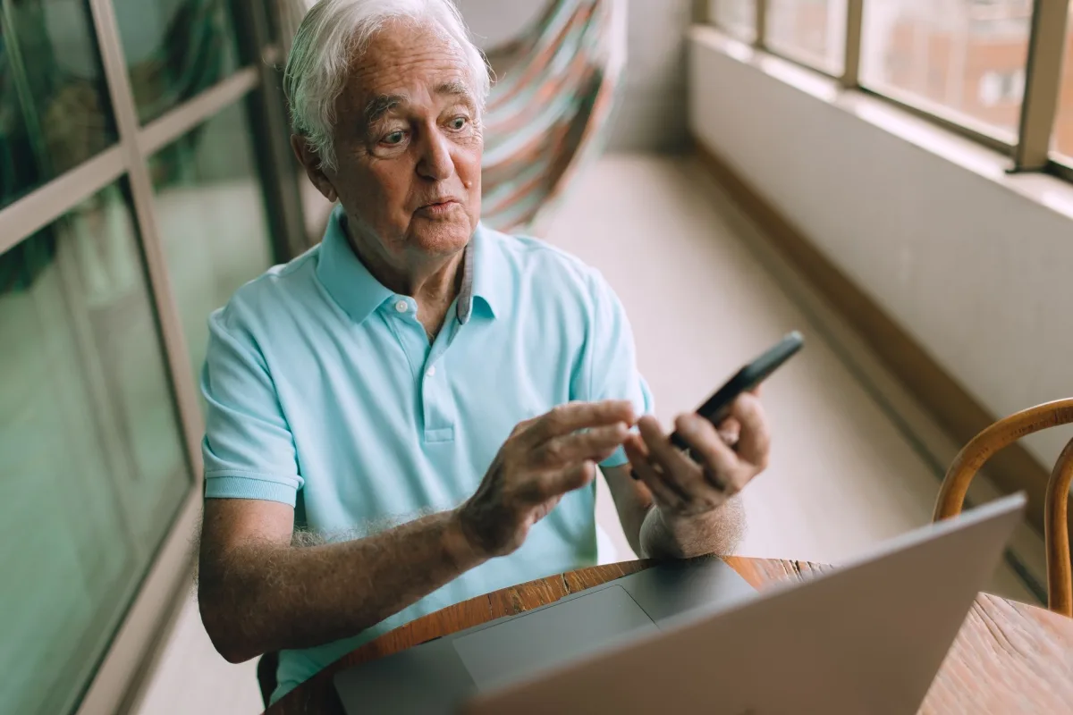 Elderly man sitting at a table, using a smartphone with a laptop nearby in an indoor setting.
