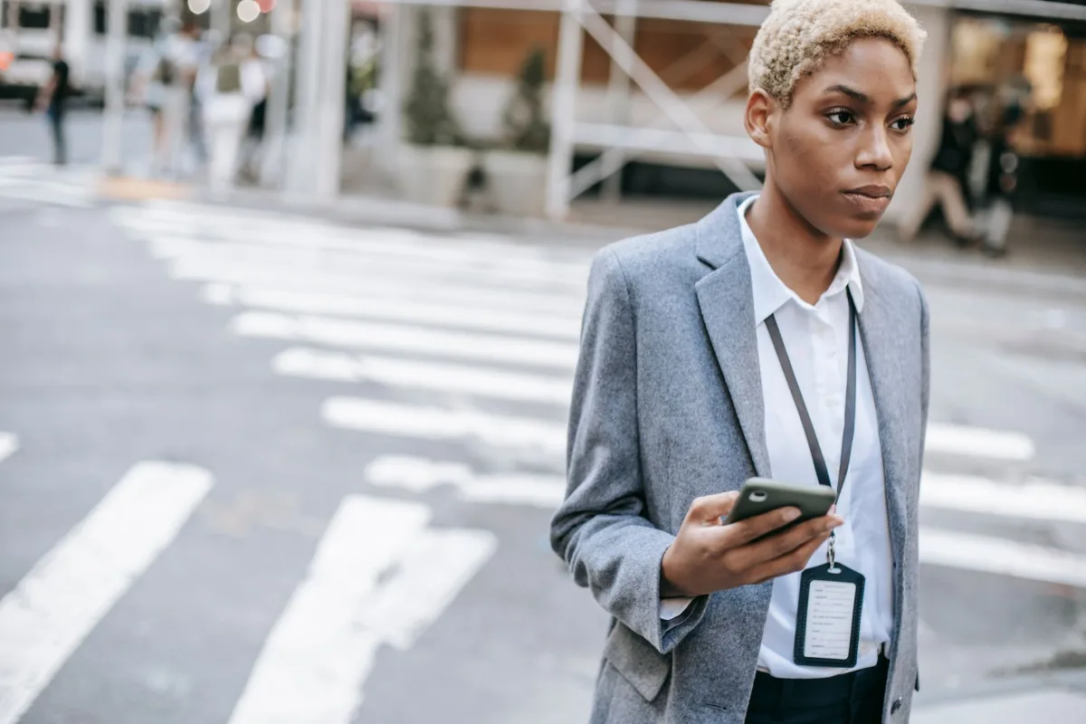 Crop young African American female entrepreneur in trendy gray coat with badge chatting on smartphone while standing on pavement near road Crop young African American female entrepreneur in trendy gray coat with badge chatting on smartphone while standing on pavement near road