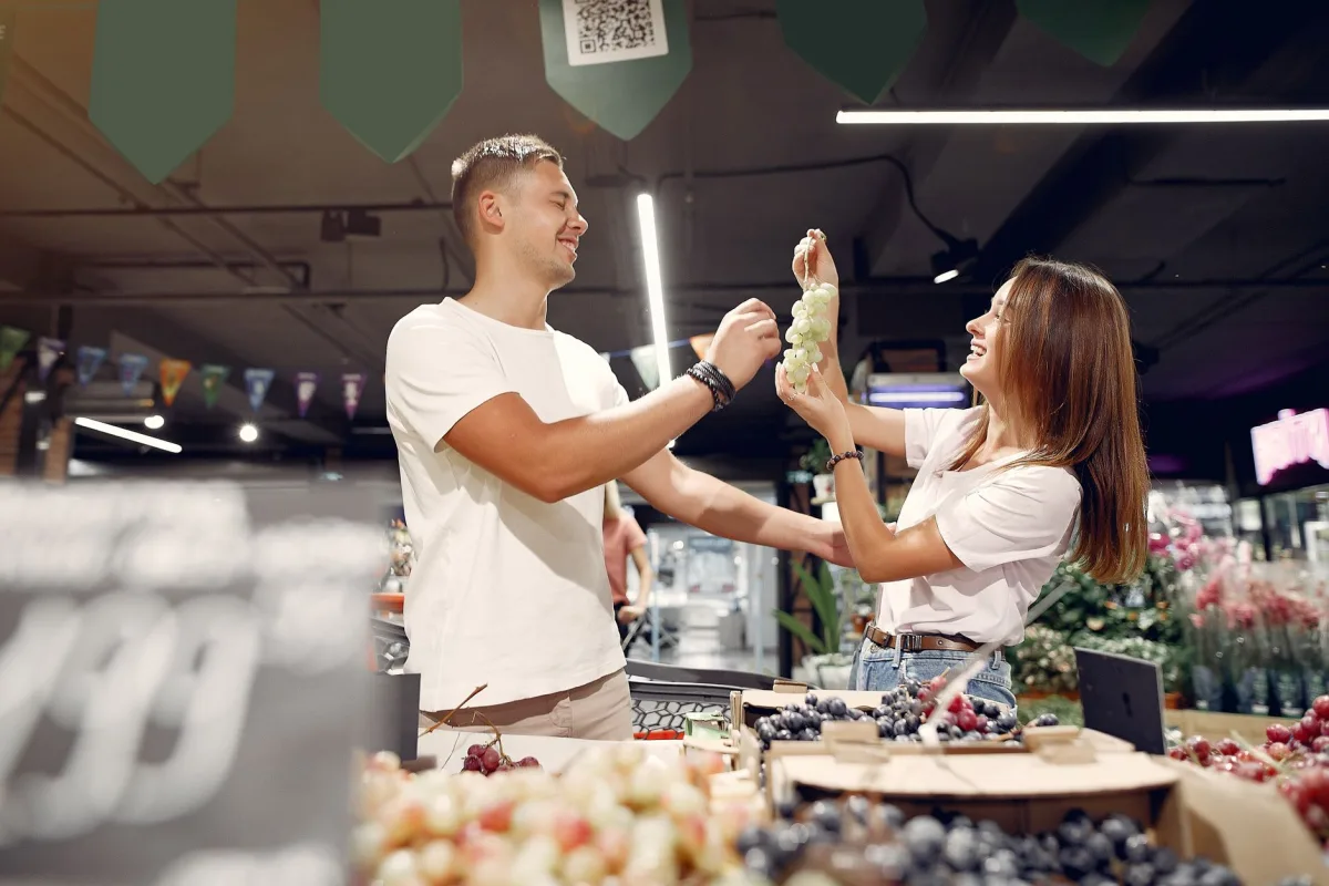 Side view of young man and woman in casual wear standing near counter and playing with each other while choosing grape in supermarket