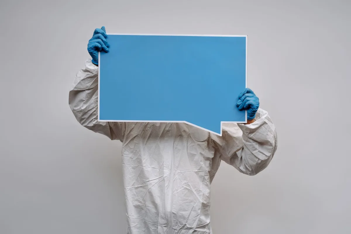 An individual in protective gear holds a blue empty speech bubble sign against a white background. An individual in protective gear holds a blue empty speech bubble sign against a white background.