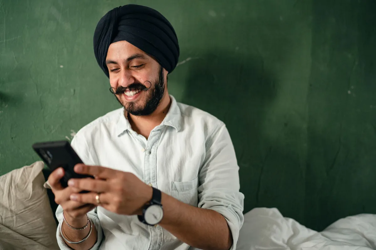 Young Indian male with black beard and mustache casual shirt and turban messaging on smartphone and smiling happily while sitting on bed near green wall at home