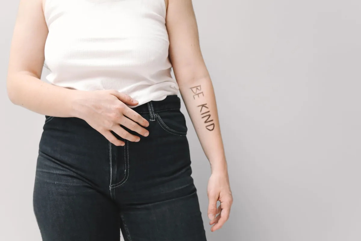 Person in jeans and white tank top with 'Be Kind' text on arm, promoting kindness.