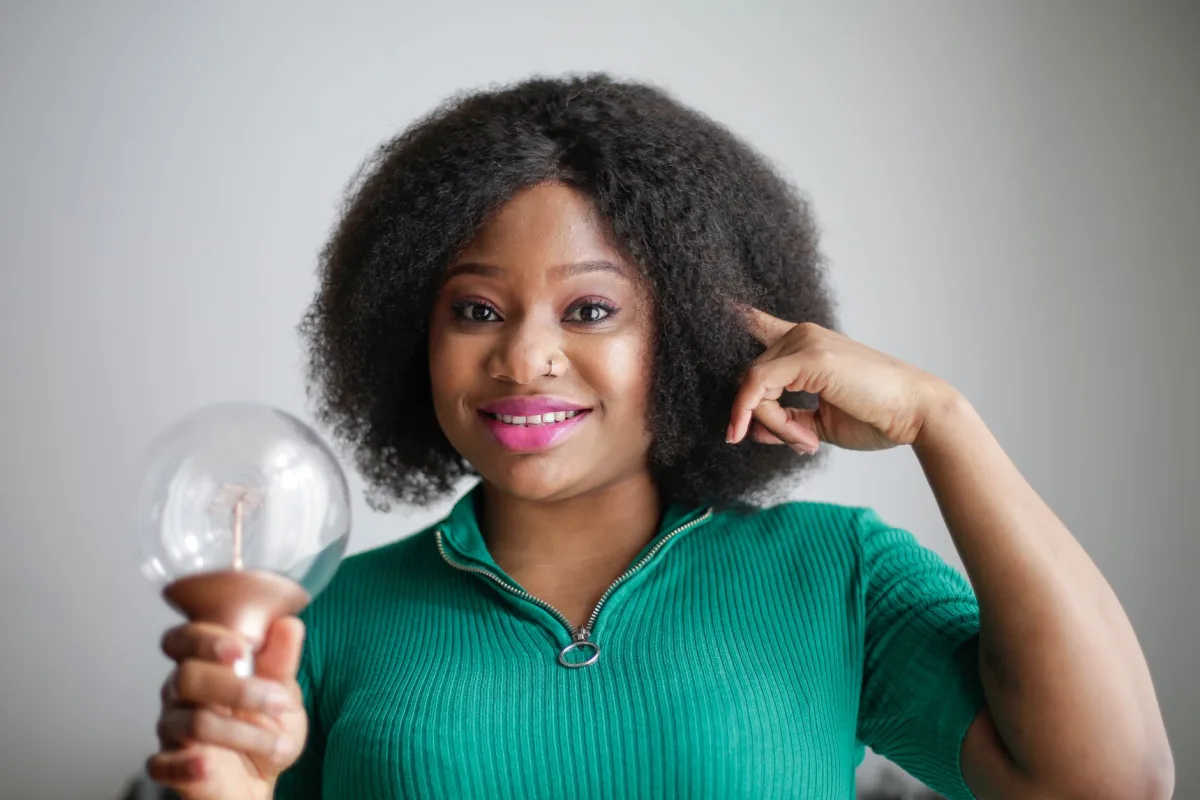 Smiling young African American lady in green casual clothes holding light bulb in hand and touching temple while having brilliant idea and looking at camera on gray background Smiling young African American lady in green casual clothes holding light bulb in hand and touching temple while having brilliant idea and looking at camera on gray background