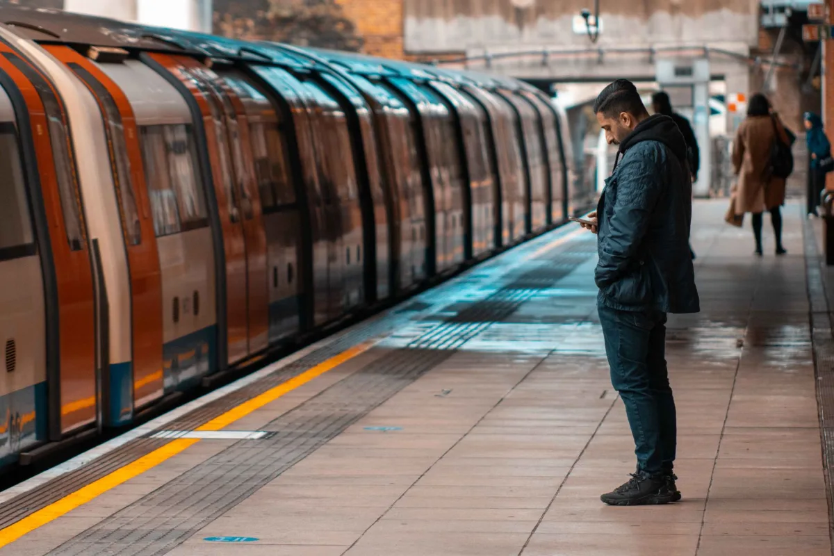 A man waits at a railway platform, texting on his cellphone, as a train arrives during daylight.