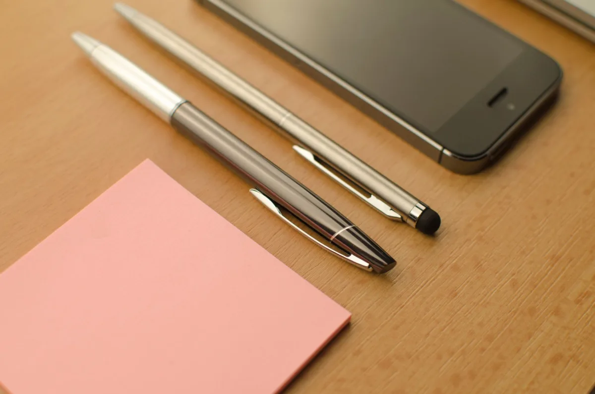 Stylish workspace featuring a smartphone, pens, and a notepad on a wooden desk. Stylish workspace featuring a smartphone, pens, and a notepad on a wooden desk.