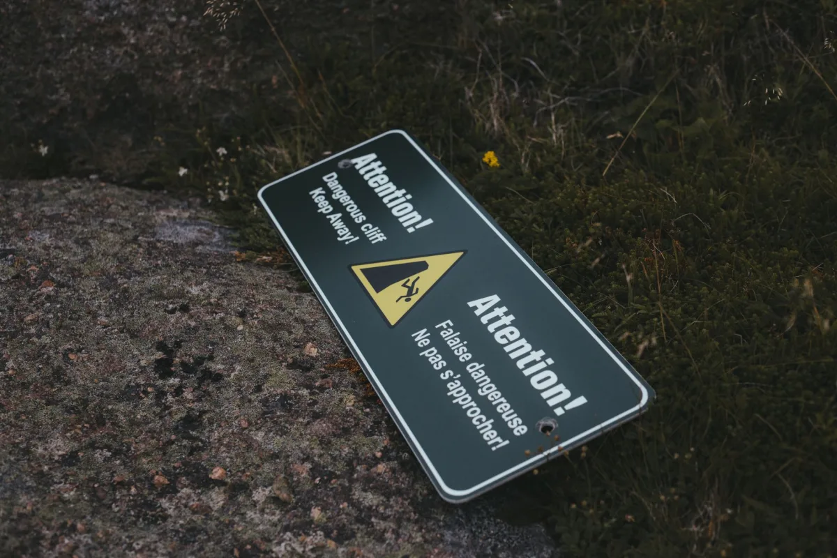 An outdoor warning sign saying 'Attention! Dangerous cliff. Keep away.' lying on rocky ground. An outdoor warning sign saying 'Attention! Dangerous cliff. Keep away.' lying on rocky ground.