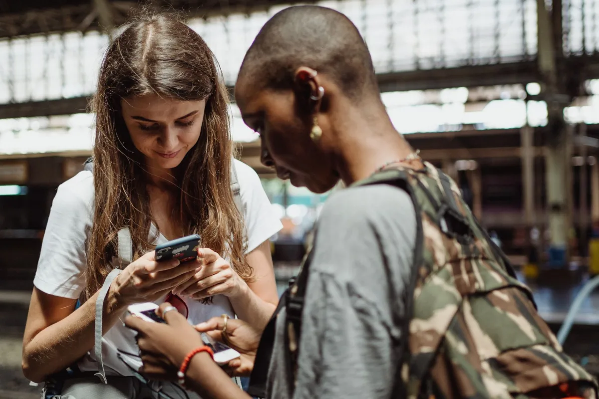 Two women using smartphones while standing in a busy train station, engaging in travel planning. Two women using smartphones while standing in a busy train station, engaging in travel planning.