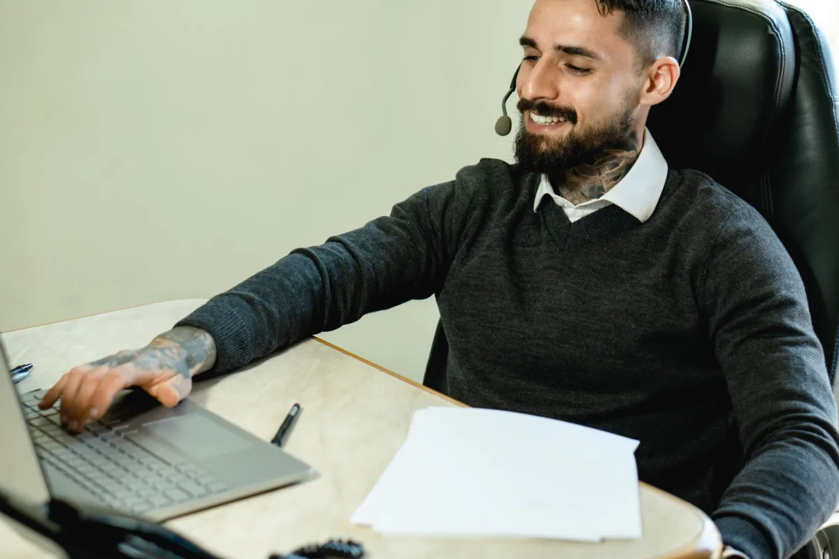 A young tattooed man in a headset works at a desk with a laptop and documents. A young tattooed man in a headset works at a desk with a laptop and documents.