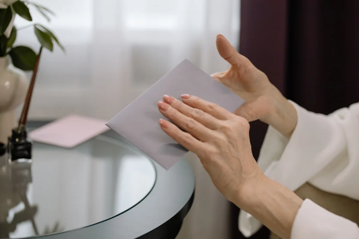 Close-up of hands holding an envelope on a glass table indoors, with a dimly lit room. Close-up of hands holding an envelope on a glass table indoors, with a dimly lit room.