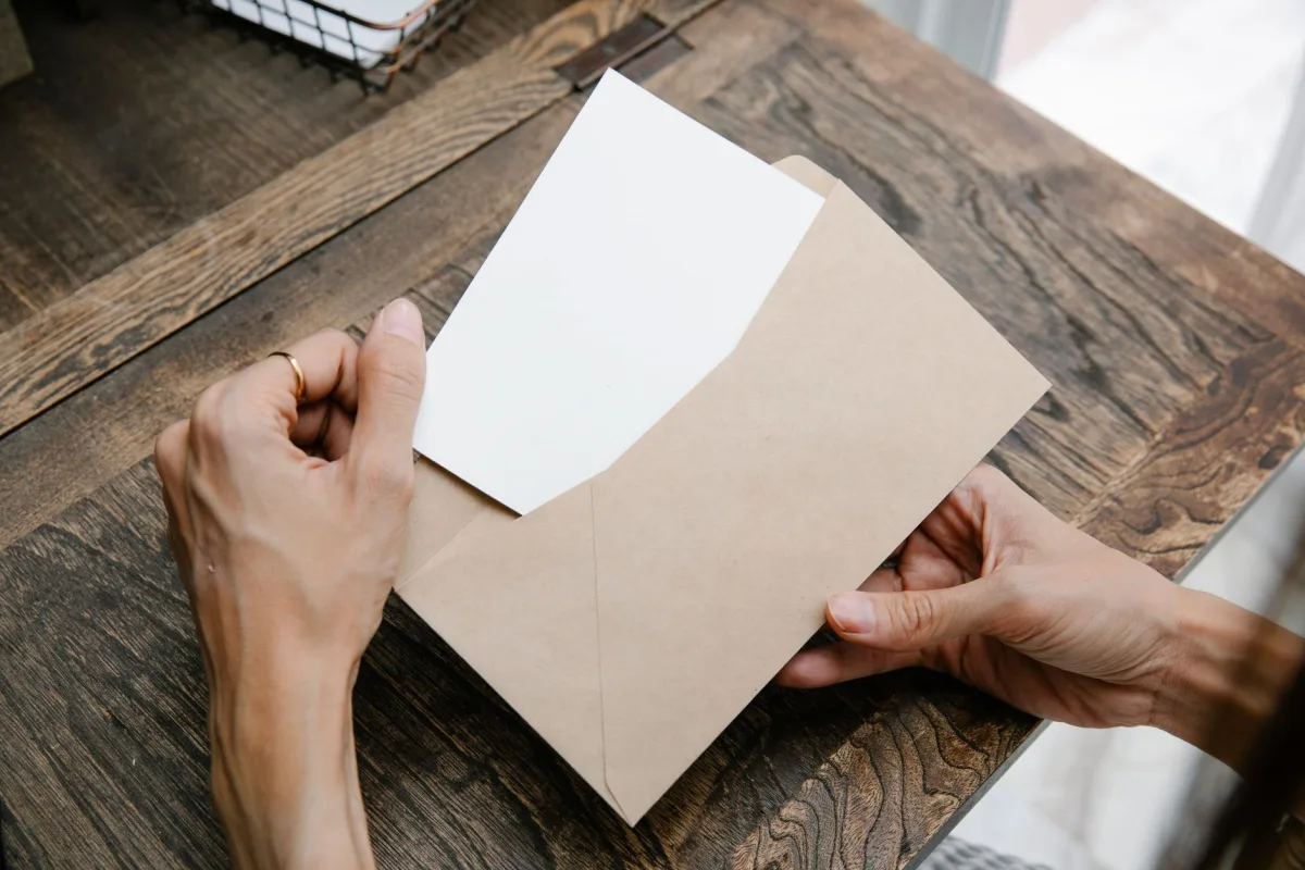 Hands holding a letter inside a brown envelope on a wooden desk, viewed from above. Hands holding a letter inside a brown envelope on a wooden desk, viewed from above.