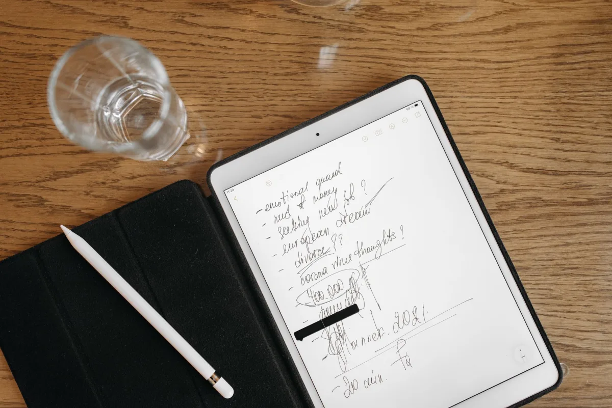 Top-down view of a tablet with handwritten notes, stylus, and glass on a wooden surface. Top-down view of a tablet with handwritten notes, stylus, and glass on a wooden surface.