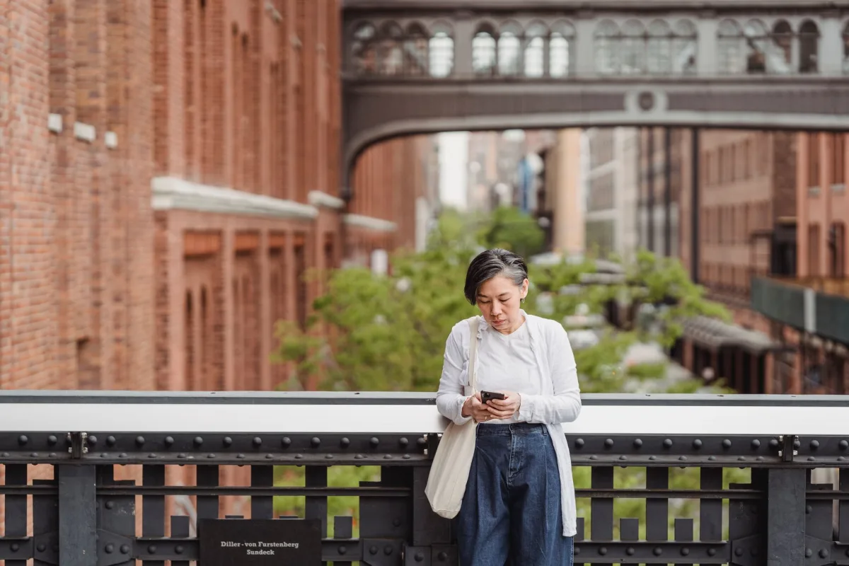 A woman uses her smartphone while leaning on a bridge in an urban setting, depicting connectivity and modern lifestyle. A woman uses her smartphone while leaning on a bridge in an urban setting, depicting connectivity and modern lifestyle.
