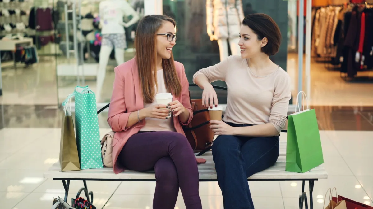 Two women chatting over coffee in a shopping mall with colorful bags.