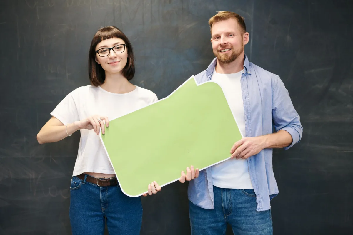 Man and woman with glasses holding a green speech bubble in front of chalkboard. Man and woman with glasses holding a green speech bubble in front of chalkboard.