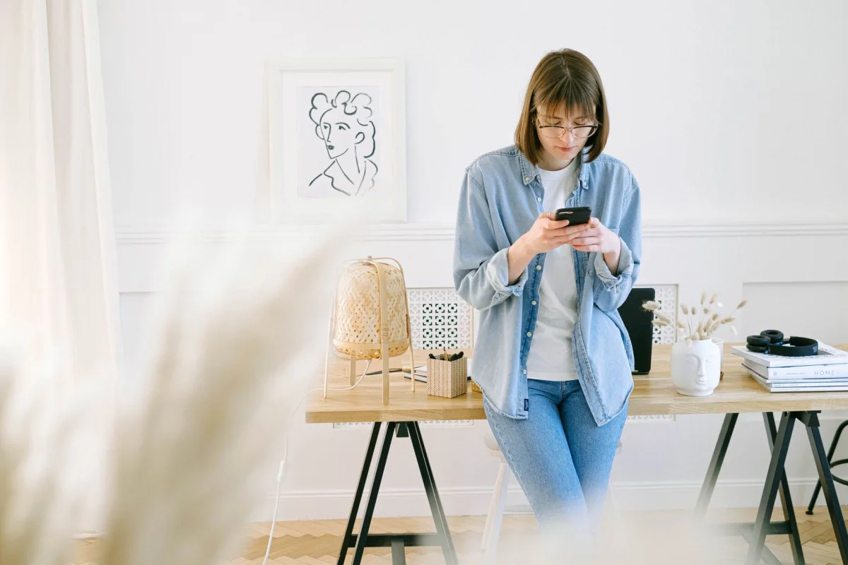 Woman in denim using smartphone in a bright home office setting.