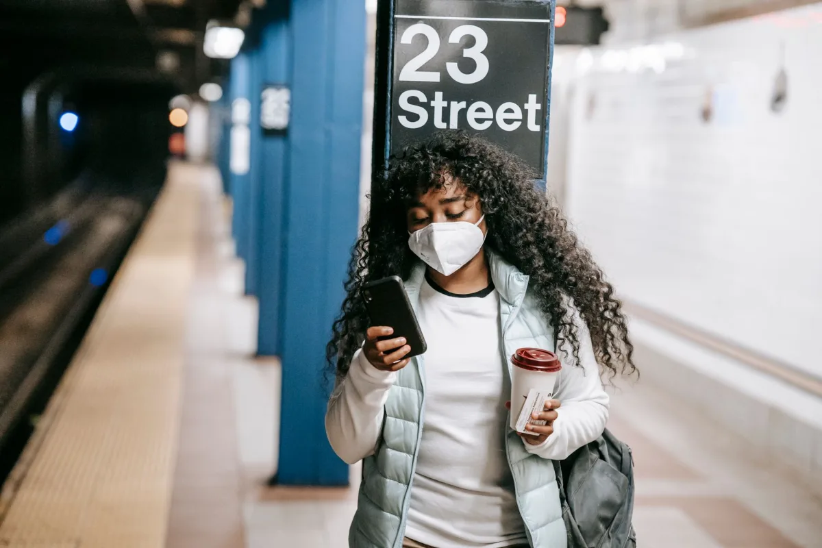 Unemotional African American female in casual clothes and respirator with takeaway coffee browsing mobile phone while waiting for train on underground platform in New York Unemotional African American female in casual clothes and respirator with takeaway coffee browsing mobile phone while waiting for train on underground platform in New York