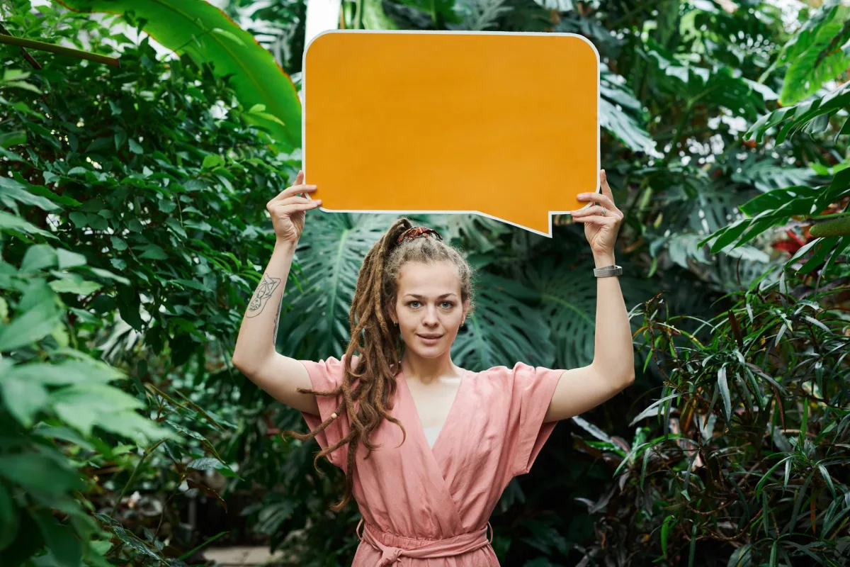 Woman with dreadlocks holding a blank sign amidst vibrant greenery, suggesting nature communication Woman with dreadlocks holding a blank sign amidst vibrant greenery, suggesting nature communication