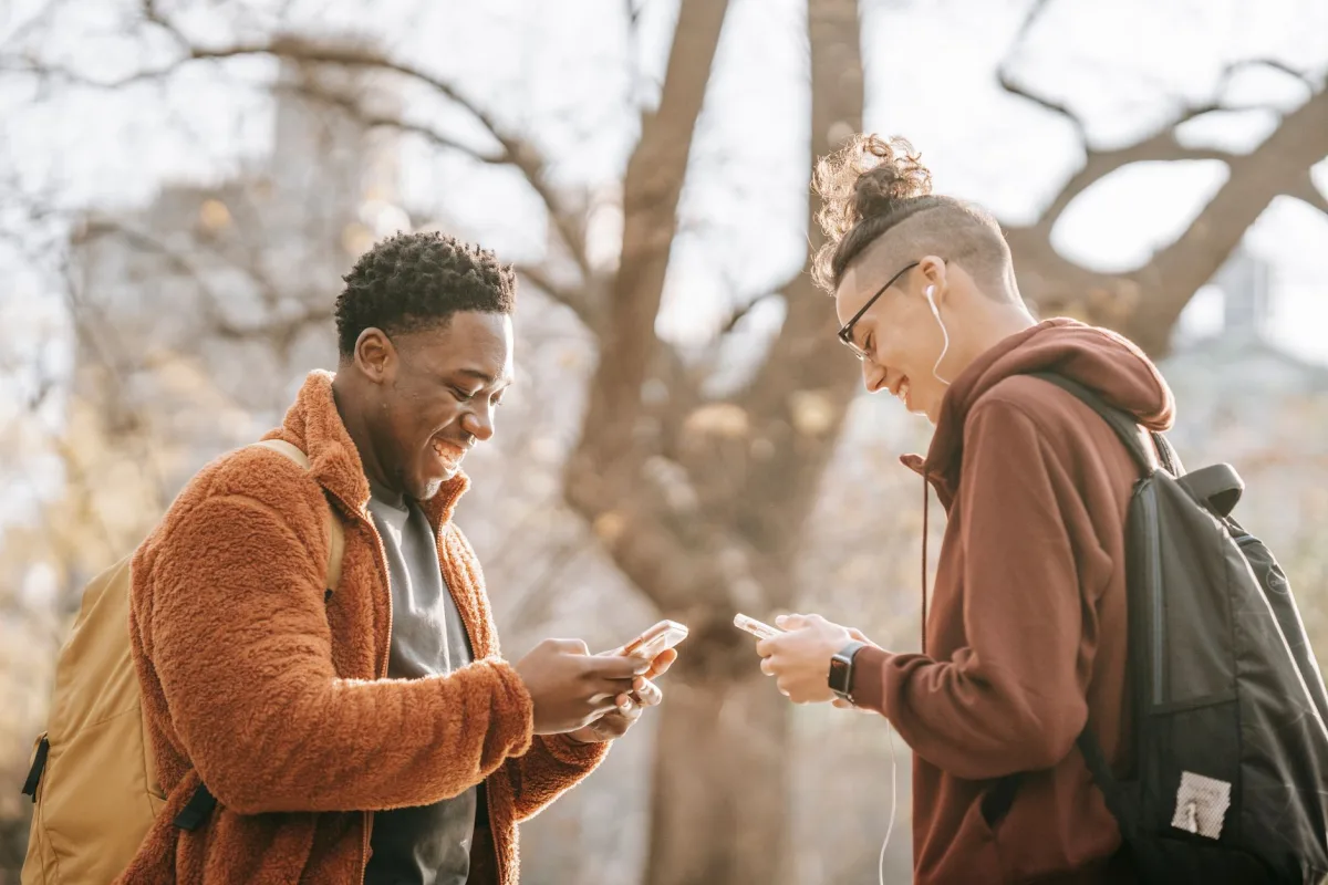 Side view of positive young diverse male friends with backpacks using contemporary cellphones while standing in city street on autumn day