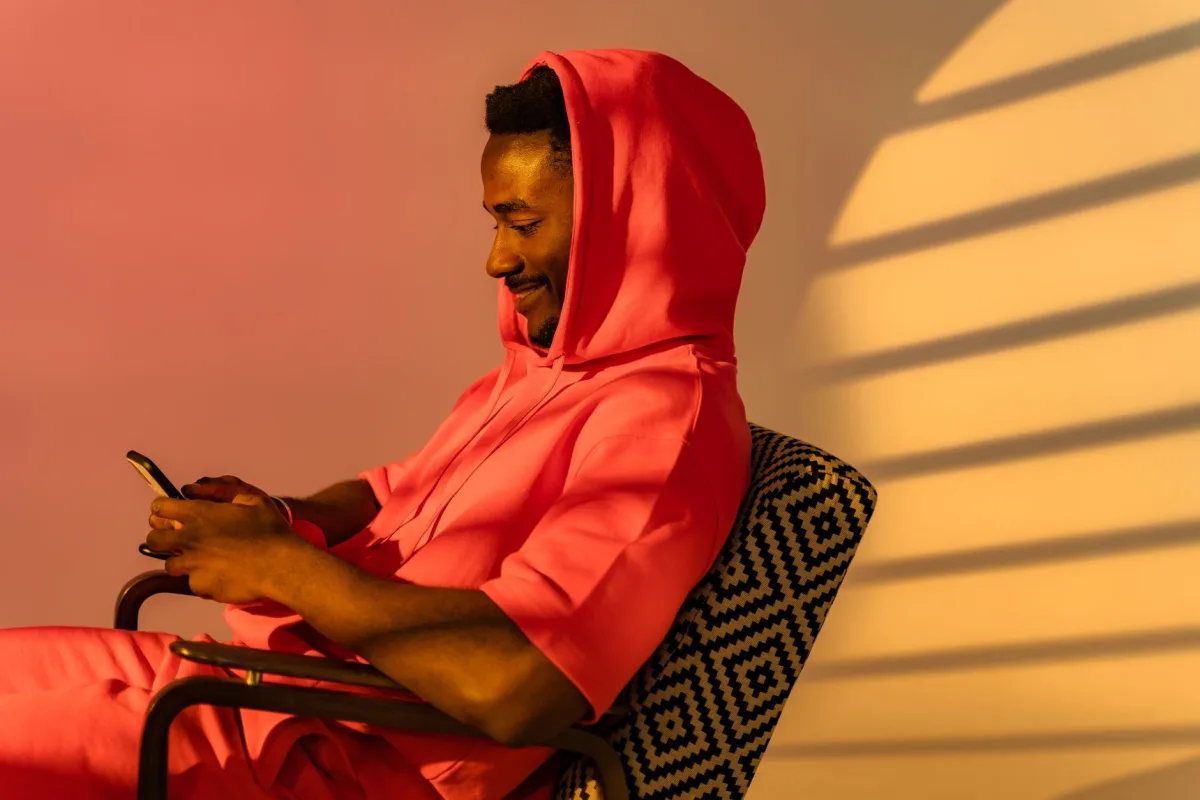 A man in a pink hoodie relaxes on a patterned chair, texting on his smartphone indoors.