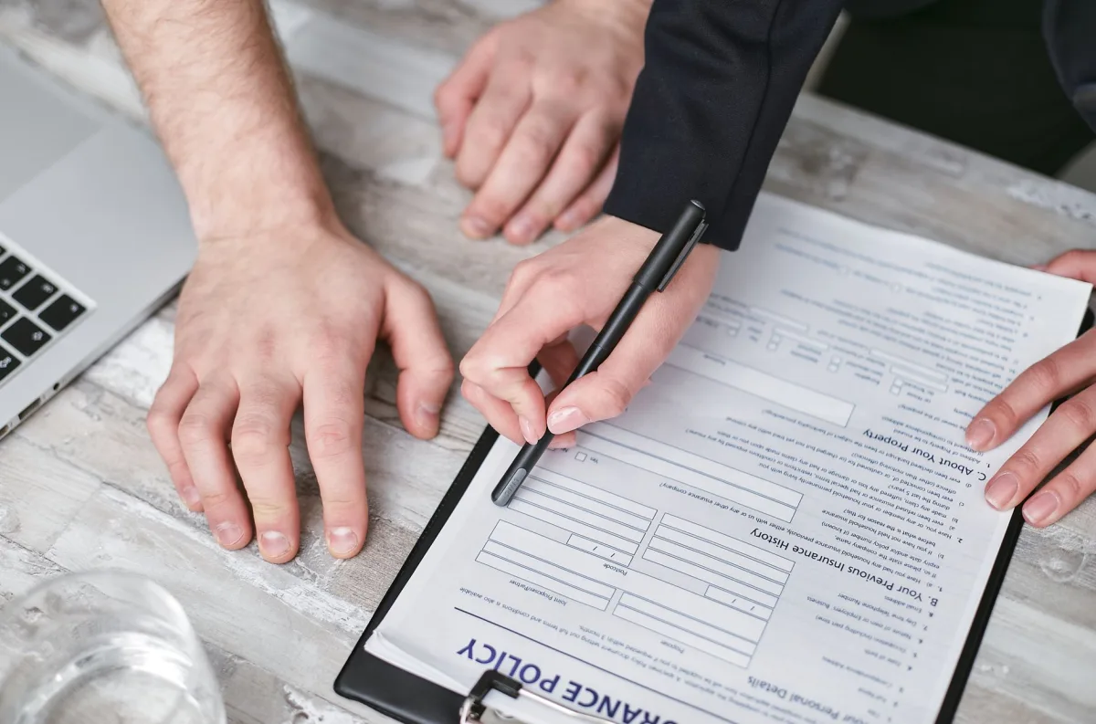 Close-up image of two people signing an insurance policy document on a wooden desk. Close-up image of two people signing an insurance policy document on a wooden desk.