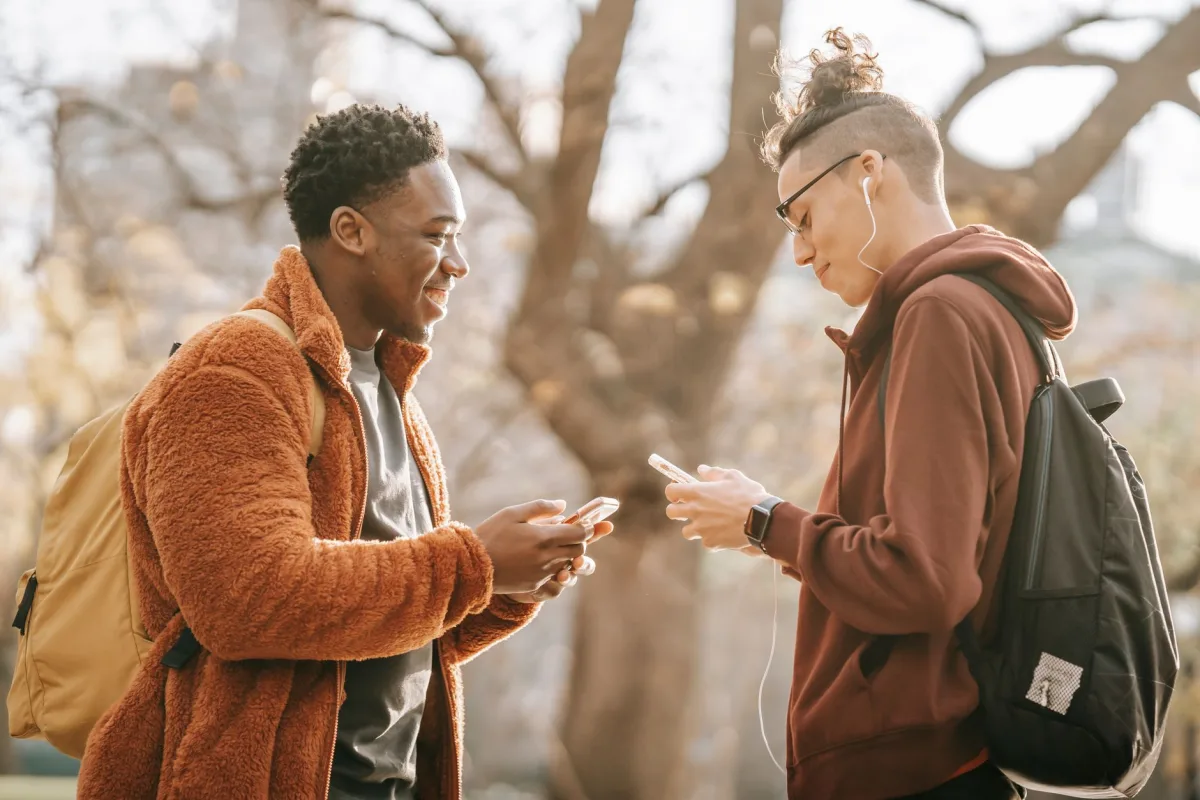Side view of smiling young multiracial male friends wearing warm clothing standing opposite each other and browsing cellphones on blurred background