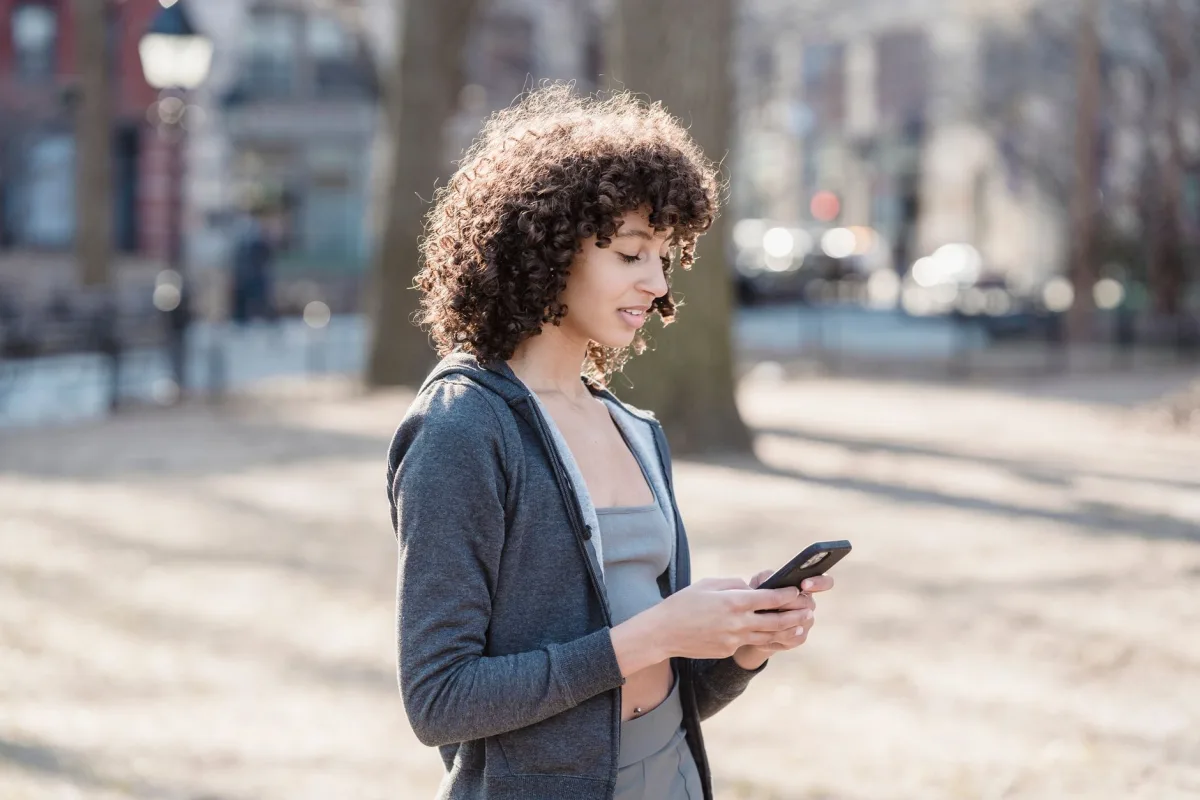 African American female in gray outfit typing text message while walking in park against blurred background African American female in gray outfit typing text message while walking in park against blurred background