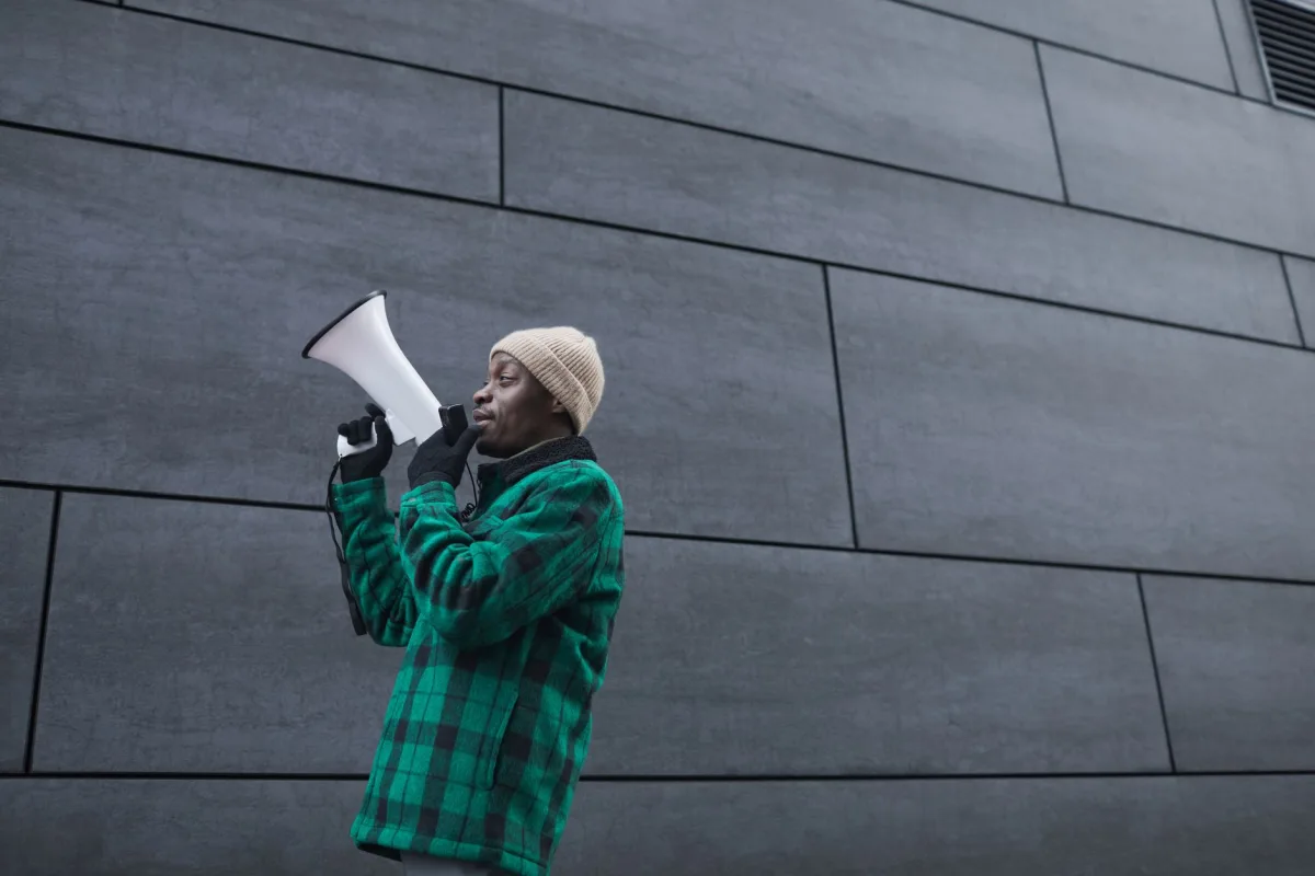 Man in green jacket speaking through a megaphone against a concrete wall backdrop.