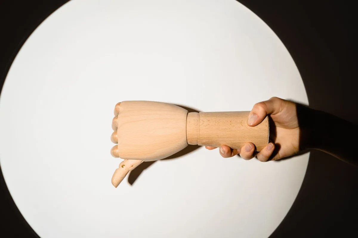 A wooden mannequin hand held by a person under a spotlight, creating a dramatic shadow effect.