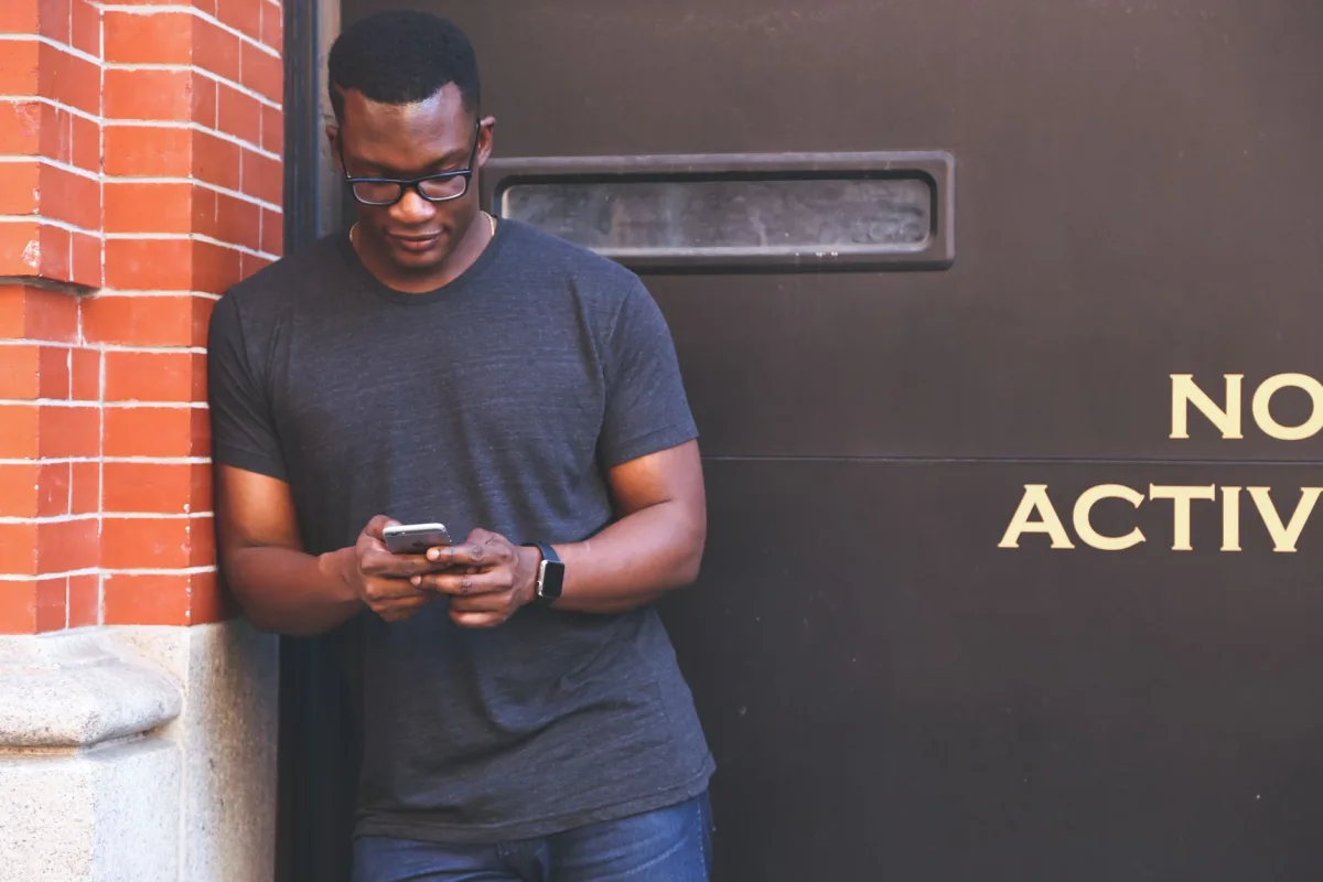 African American man using smartphone while leaning against a brick wall, wearing casual attire.