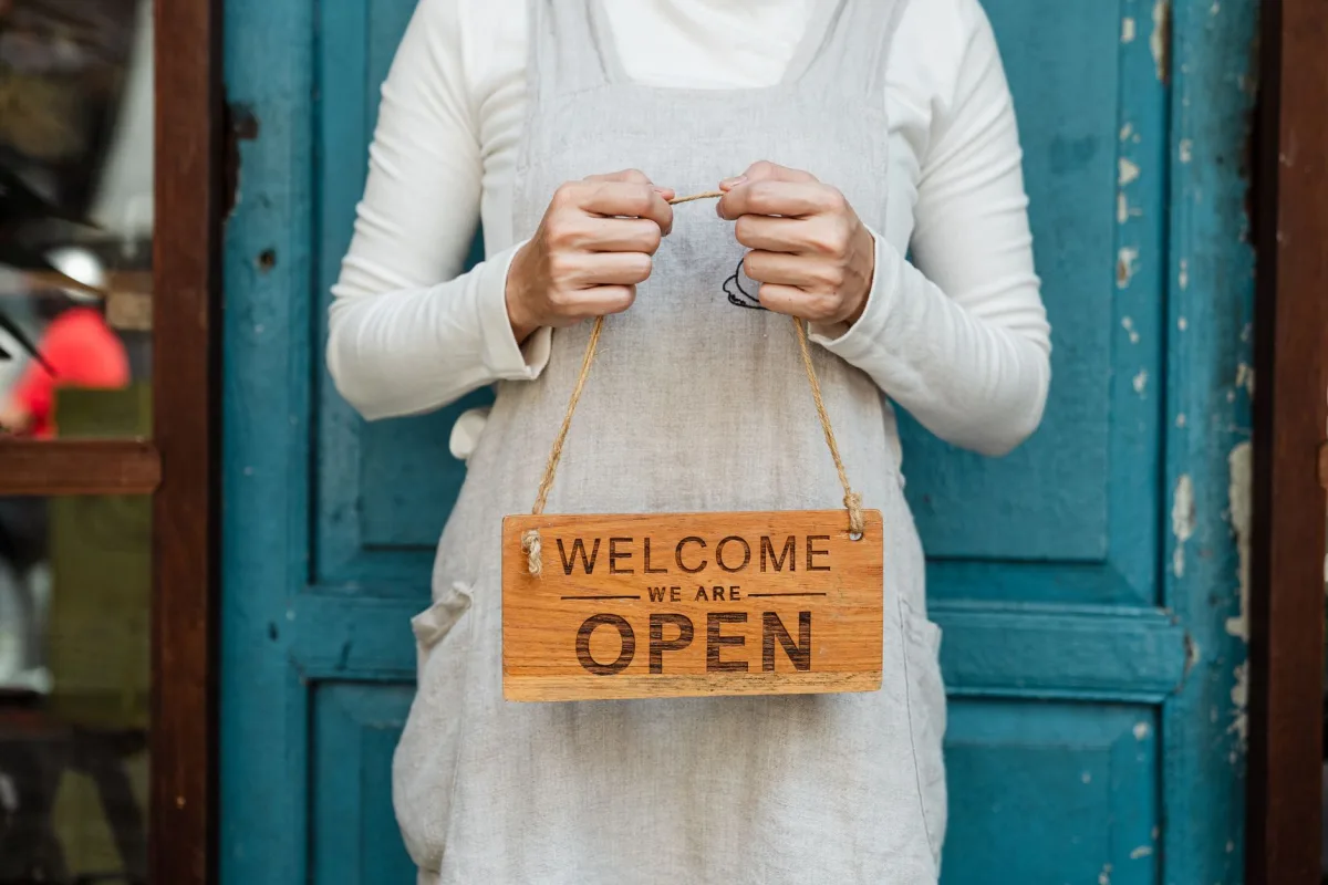 A person holds a "Welcome, we are open" sign in front of a rustic blue door. A person holds a "Welcome, we are open" sign in front of a rustic blue door.