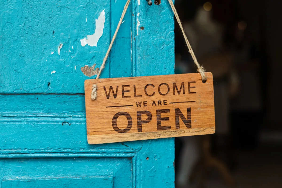 A rustic wooden 'Welcome We Are Open' sign hanging on a vibrant blue door. A rustic wooden 'Welcome We Are Open' sign hanging on a vibrant blue door.