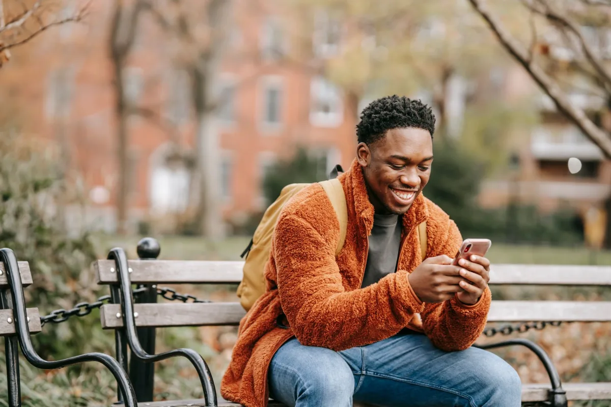 Positive young African American guy wearing warm clothes sitting on wooden bench in urban park and chatting on mobile phone on autumn day