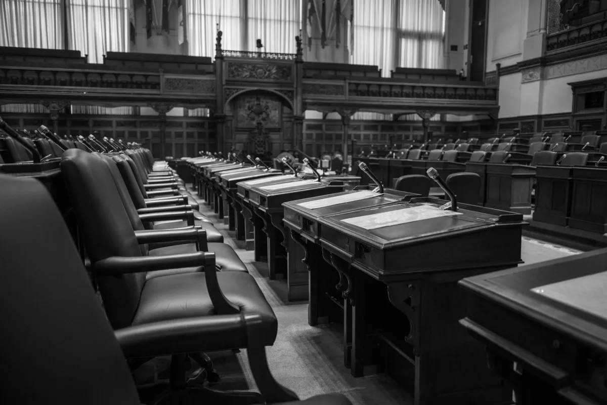 Black and white photo of a traditional legislative assembly chamber, featuring empty seats and desks. Black and white photo of a traditional legislative assembly chamber, featuring empty seats and desks.