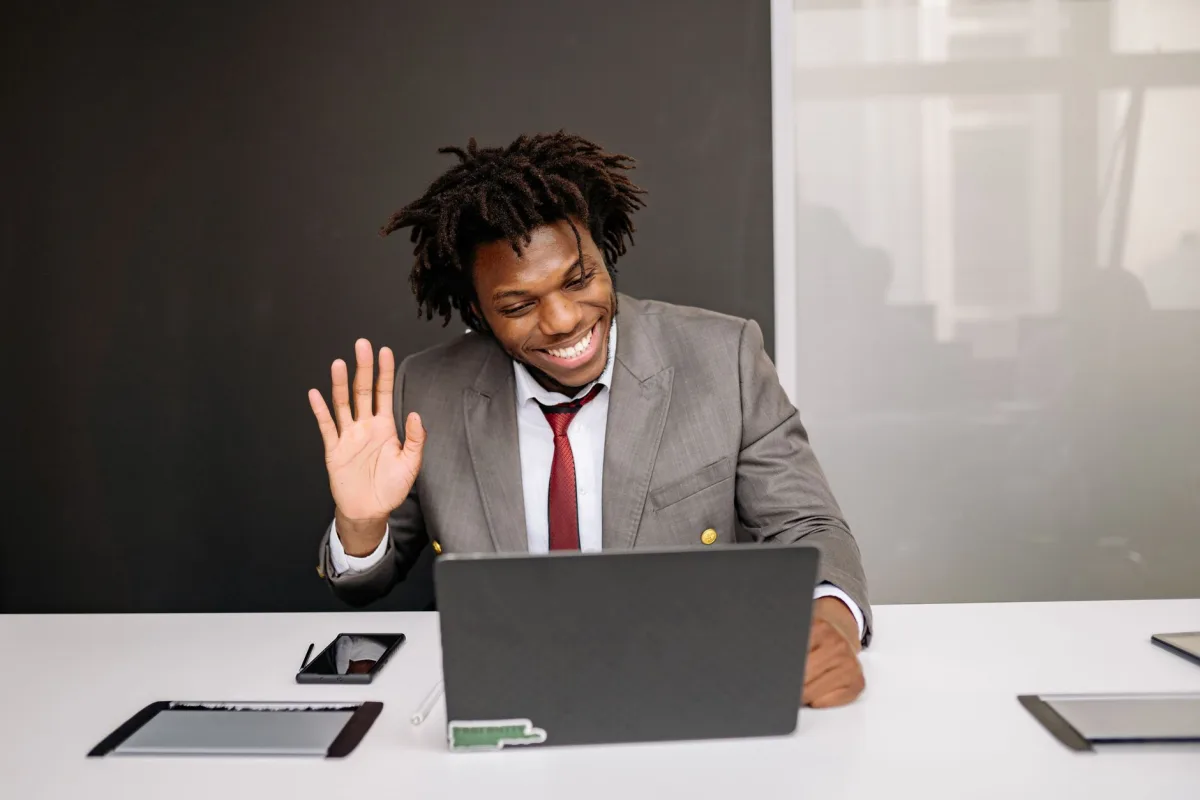 Professional making a video call, waving hand at laptop in modern office setting. Professional making a video call, waving hand at laptop in modern office setting.