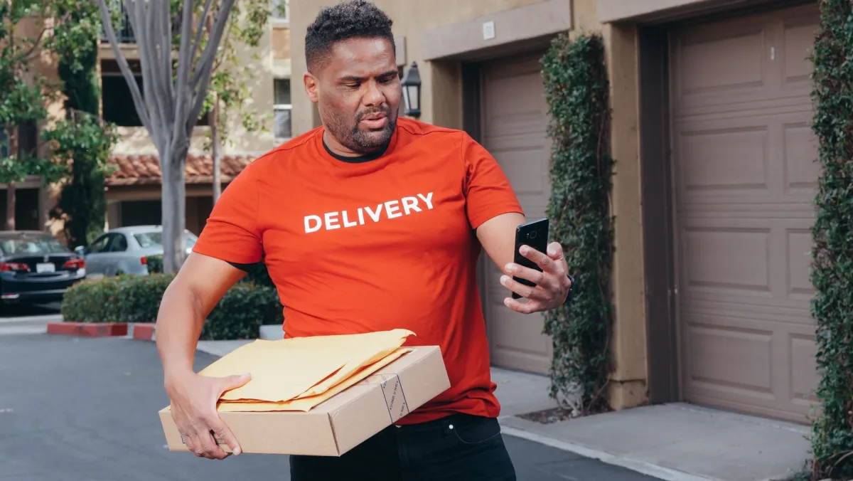 A delivery man looking at his smartphone while holding packages, outdoors. A delivery man looking at his smartphone while holding packages, outdoors.