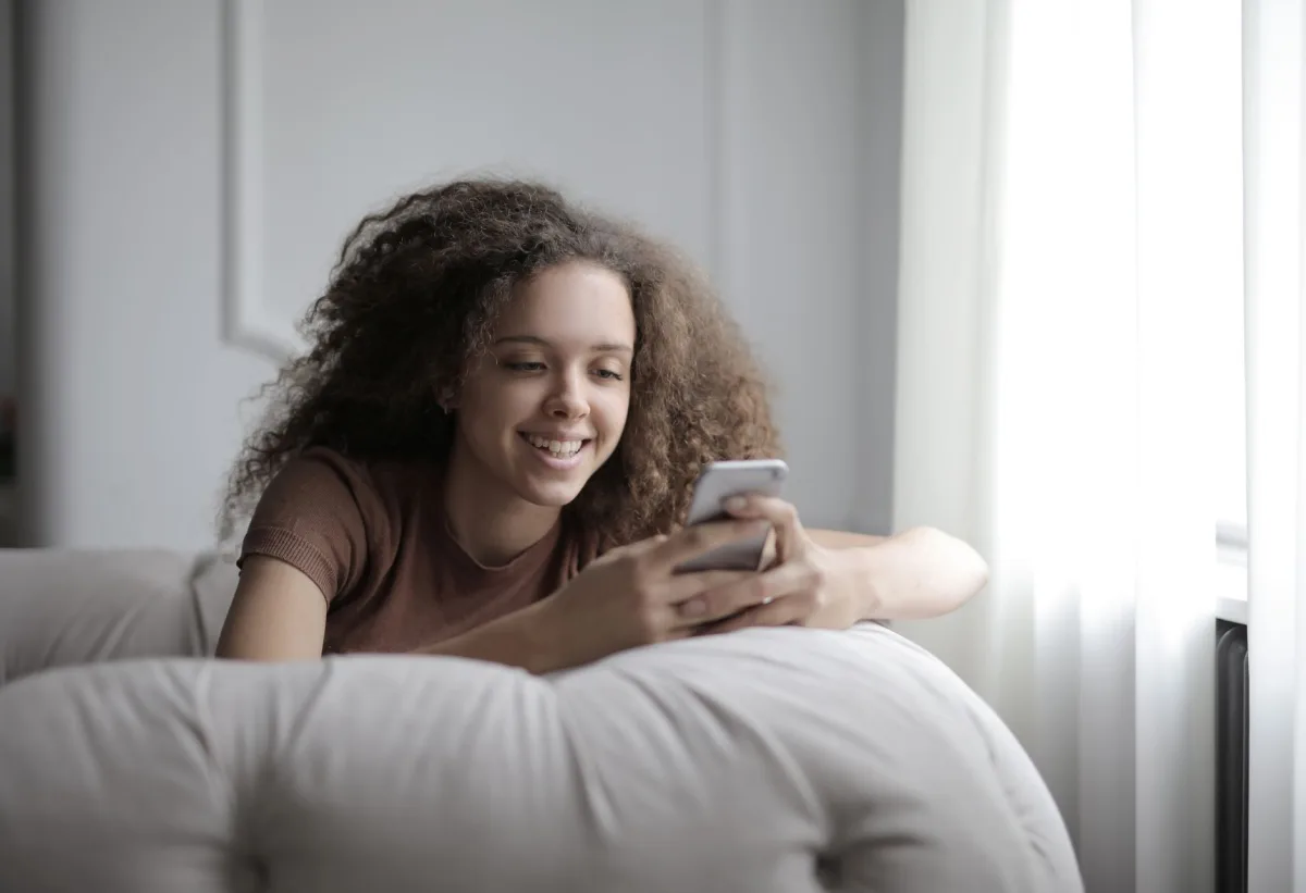Curly teenage girl in casual clothes smiling while browsing mobile phone resting on cozy sofa in living room at home Curly teenage girl in casual clothes smiling while browsing mobile phone resting on cozy sofa in living room at home
