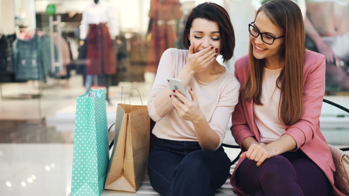 Two women sitting, laughing and looking at a phone in a shopping mall, surrounded by bags.