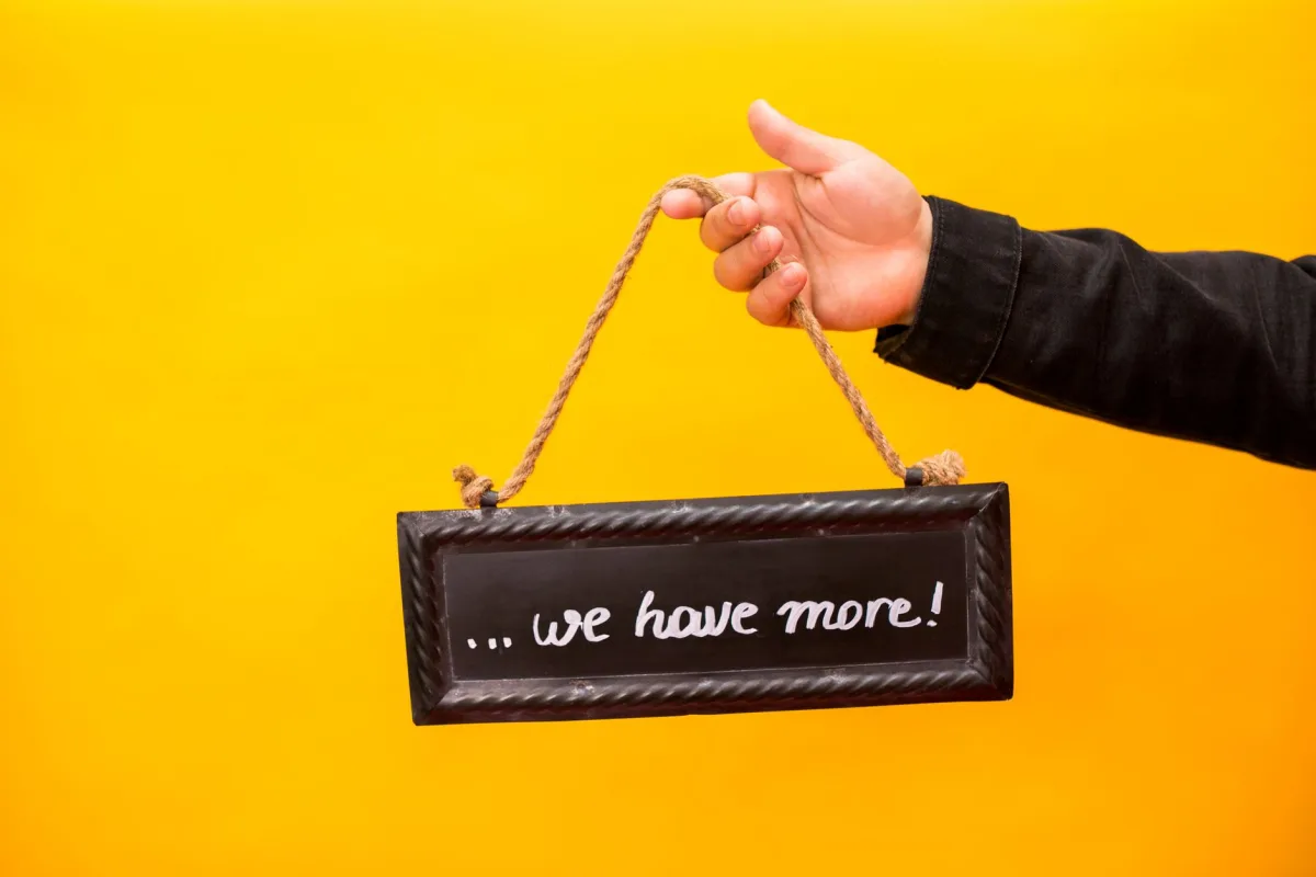 A hand holding a sign reads 'we have more' against a yellow background.