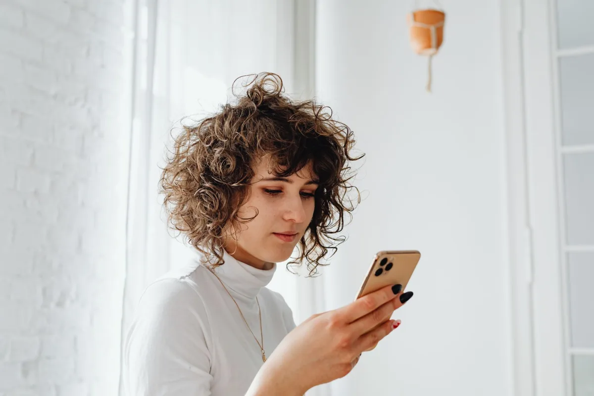 A young woman with curly hair using a smartphone indoors. Natural light and modern decor.