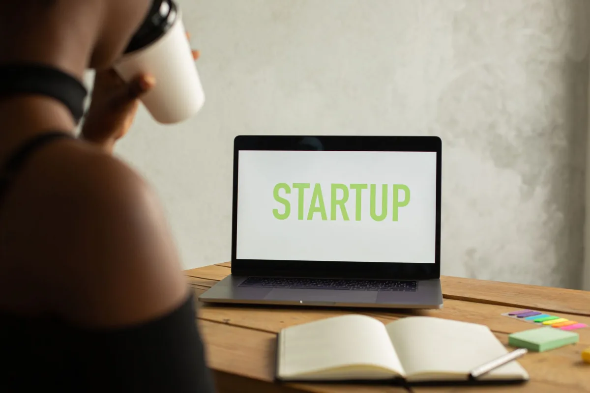 Crop anonymous black female entrepreneur sitting at table with opened netbook with startup on screen and notebook and drinking coffee from takeaway cup while working on project remotely Crop anonymous black female entrepreneur sitting at table with opened netbook with startup on screen and notebook and drinking coffee from takeaway cup while working on project remotely