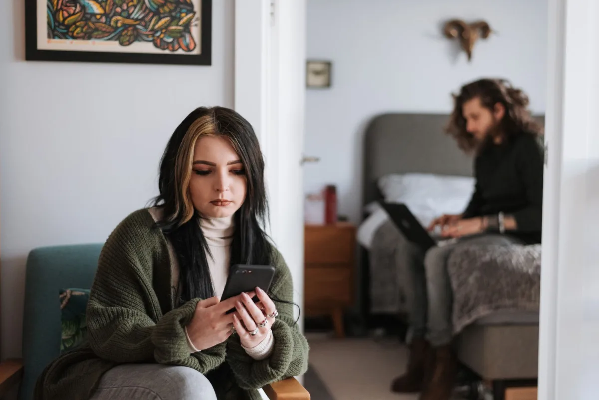 Offended young female millennial in warm clothes messaging on smartphone sitting in armchair during weekend at home with busy boyfriend working online on netbook