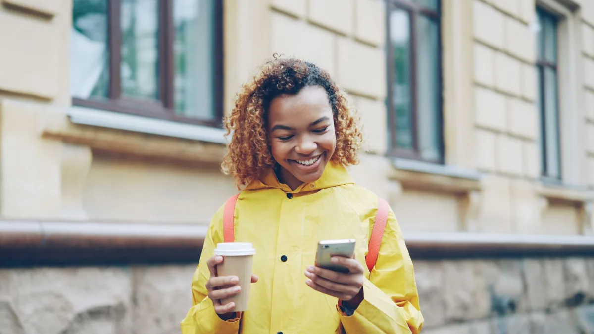 Young woman in a yellow raincoat smiling while looking at her phone and holding a coffee cup. Young woman in a yellow raincoat smiling while looking at her phone and holding a coffee cup.