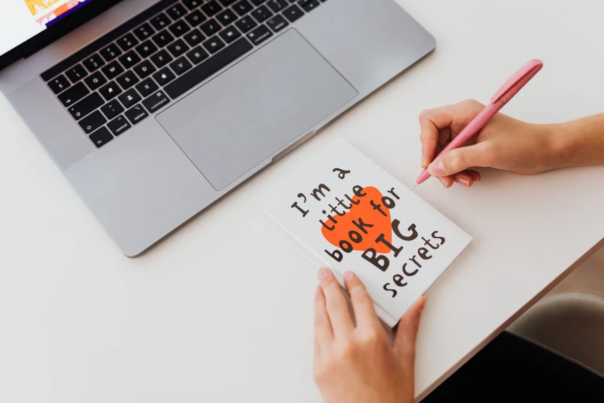 Overhead view of a woman in a notebook labeled 'I'm a little book for BIG secrets' beside a laptop.