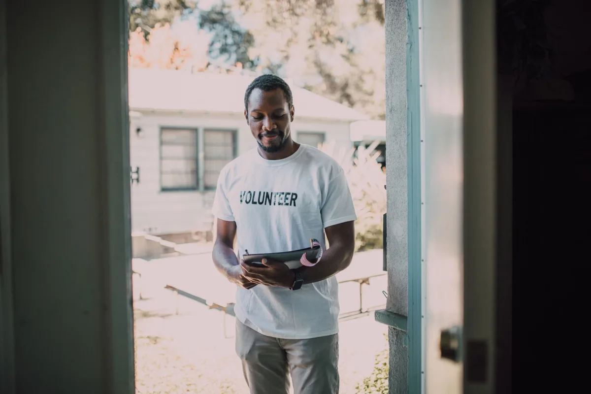 A man wearing a volunteer t-shirt stands by a doorway holding a tablet, engaged in a community event. A man wearing a volunteer t-shirt stands by a doorway holding a tablet, engaged in a community event.