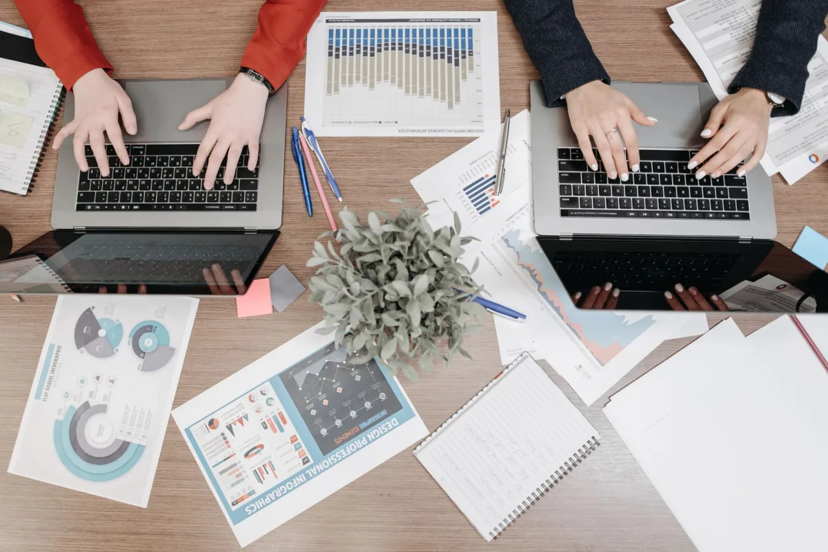 High-angle view of colleagues working on laptops with reports and charts in an office setting. High-angle view of colleagues working on laptops with reports and charts in an office setting.
