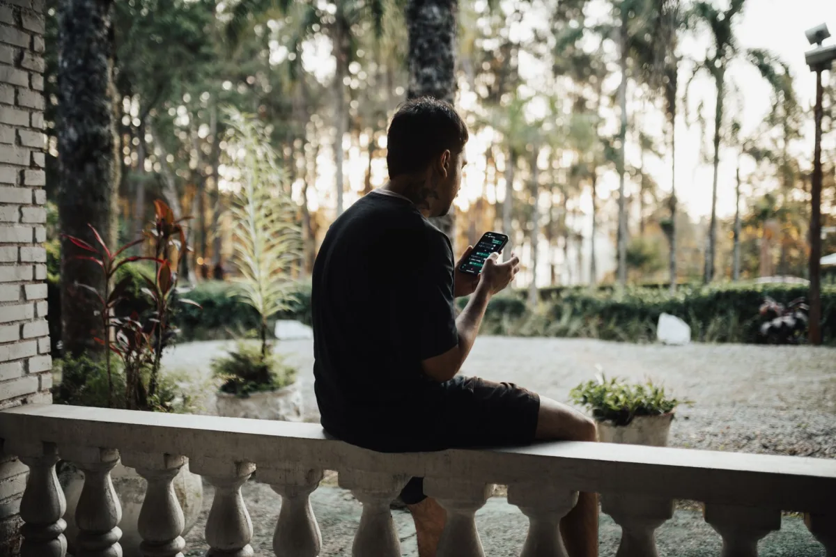 A man sitting on a patio bench his smartphone, surrounded by lush greenery and palm trees. A man sitting on a patio bench his smartphone, surrounded by lush greenery and palm trees.